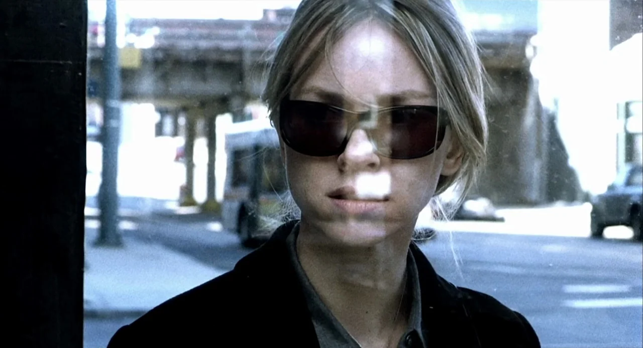 21 Grams (2003) movie still: closeup — A woman stands behind a glass pane in an urban environment with elevated tracks. She looks ahead while…