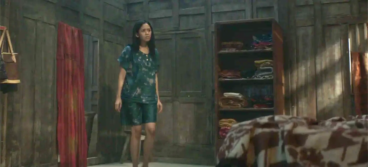 A Thousand Days (2023) movie still: medium — A dark, wood-paneled room features a small bookshelf and a wooden wall. A young woman stands in the…