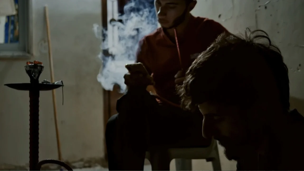 No Other Land (2024) movie still: medium — A dimly lit room with plain walls features two men in the foreground. One man checks his smartphone while…