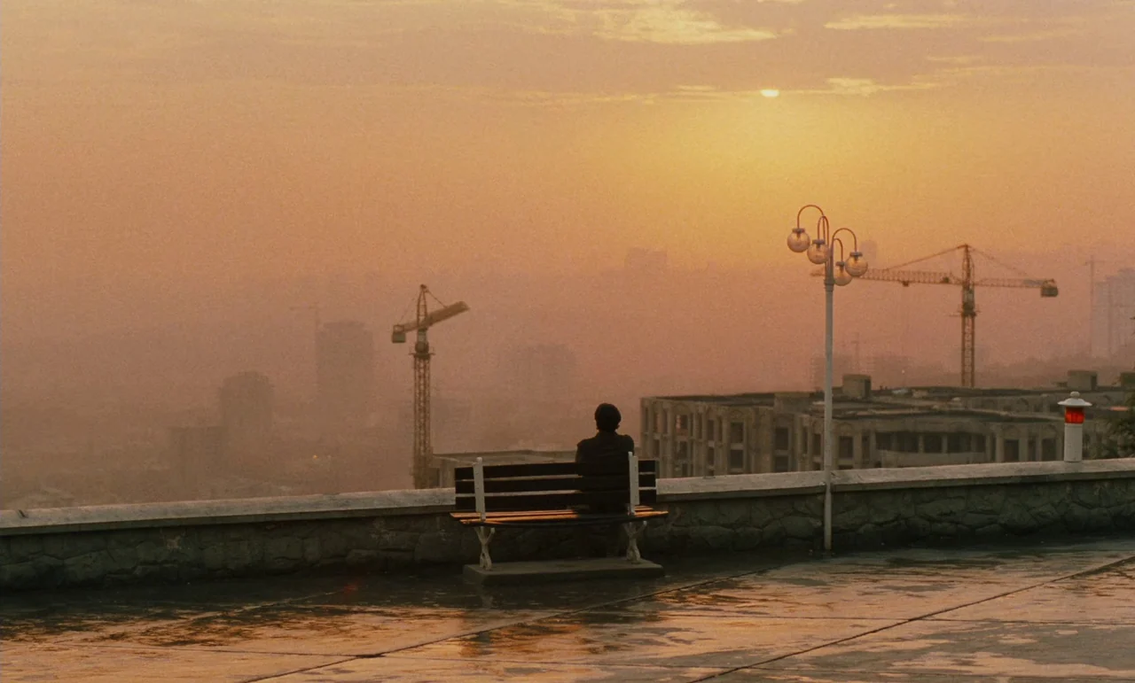 Taste of Cherry (1997) movie still: wide — A high-altitude stone overlook features a bench facing a city under a golden haze. A solitary person sits…