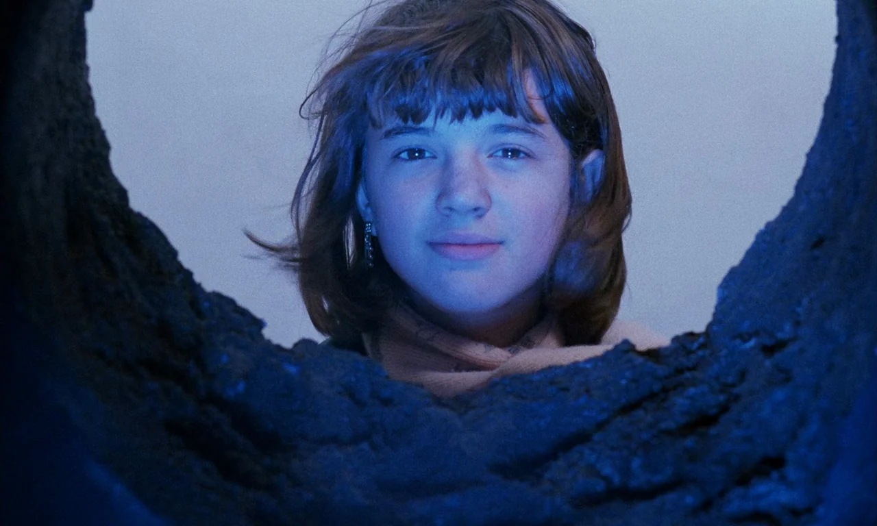 The Church (1989) movie still: closeup — A dark, uneven rock aperture frames a young girl looking out into the light. She is centered in the frame,…