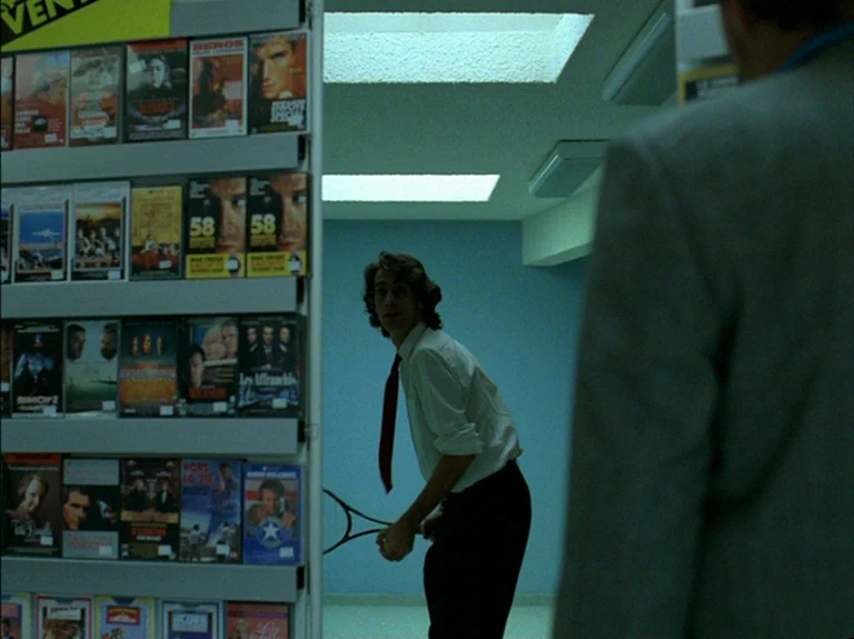 Helas Pour Moi (1993) movie still: medium — A video store aisle features shelves stocked with various VHS cases under ceiling panels. A young man in…