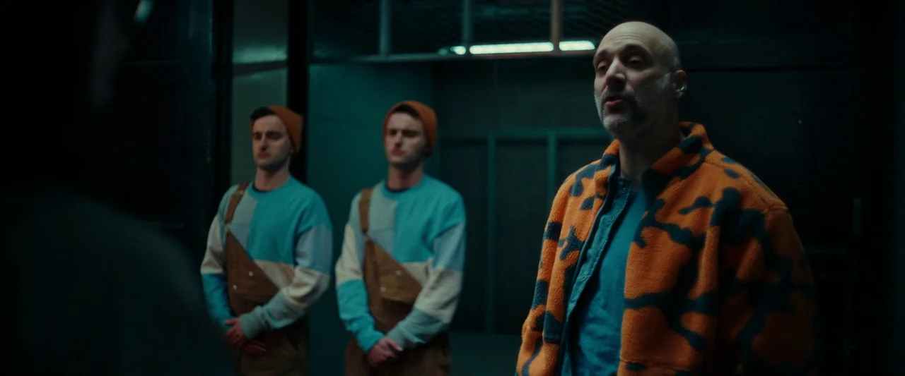 We Are Zombies (2023) movie still: medium — A dimly lit industrial space with dark walls and metallic structures visible. An older man addresses two…