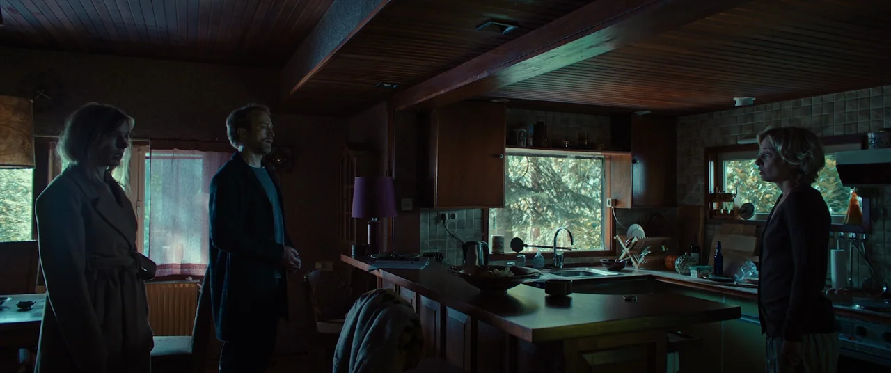 Speak No Evil (2022) movie still: wide — A wood-paneled kitchen features brown cabinetry and a breakfast bar separating the cooking area. Three…