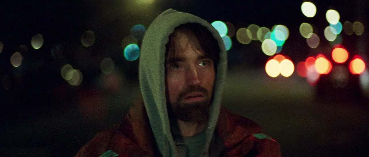 Good Time (2017) movie still: closeup — A man stands outdoors at night in front of blurred bokeh city lights. He is wearing a grey hooded sweatshirt…