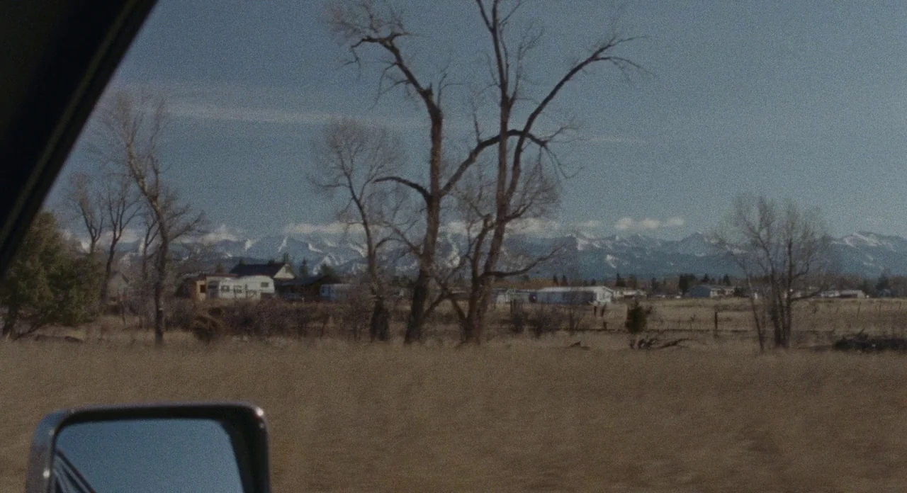 Certain Women (2016) movie still: establishing — A rural landscape features dry brown grass, bare trees, and mobile homes. Snow-capped mountains…