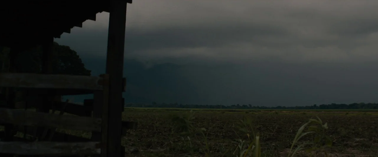 Mudbound (2017) movie still: establishing — A vast, flat, dark soil field stretches under a heavy, overcast storm sky. A wooden cattle enclosure…
