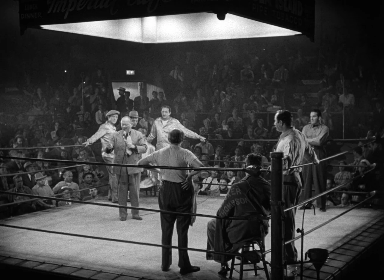 The Set-Up (1949) movie still: wide — An indoor sports venue with a brightly lit ring surrounded by a seated crowd. Several men in suits and…