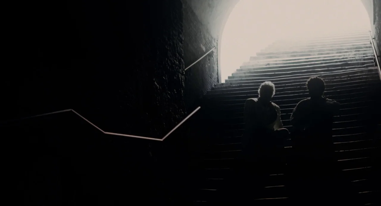 Pasolini (2014) movie still: wide — A stone stairwell with dark brick walls leads up toward a bright arched opening. Two people are silhouetted…