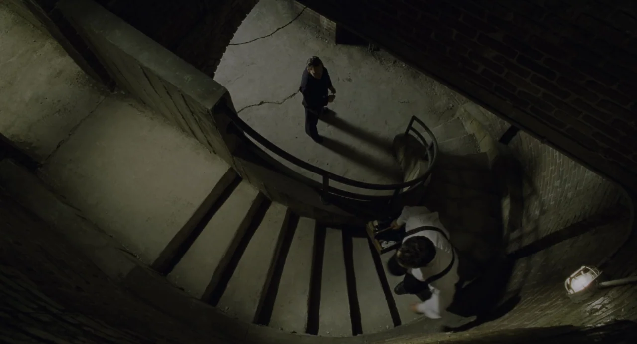 The Green Mile (1999) movie still: wide — A dark concrete spiral stairwell features exposed brick walls and a single metal railing. A man carrying a…