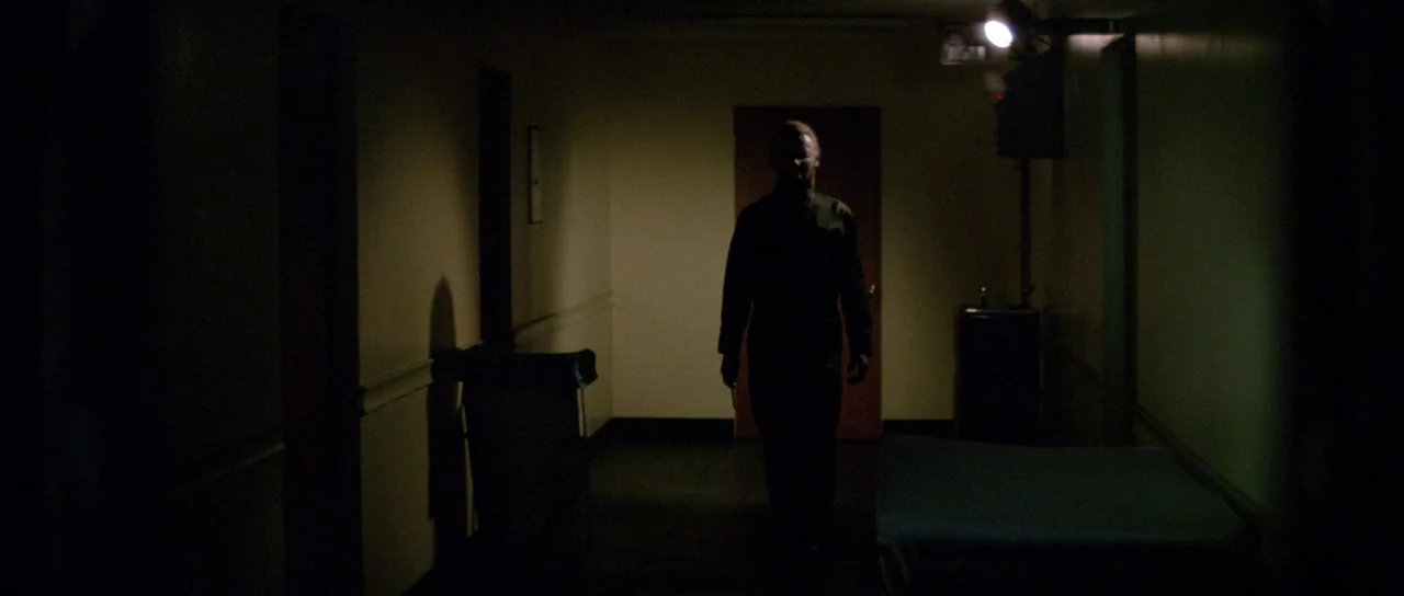 Halloween II (1981) movie still: fullbody — A dark, narrow hospital hallway has a single light fixture on the ceiling. A man walks forward toward…