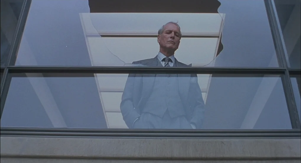 The Hudsucker Proxy (1994) movie still: medium — An elderly man stands on a balcony behind a large pane of fractured glass. The frame captures him…