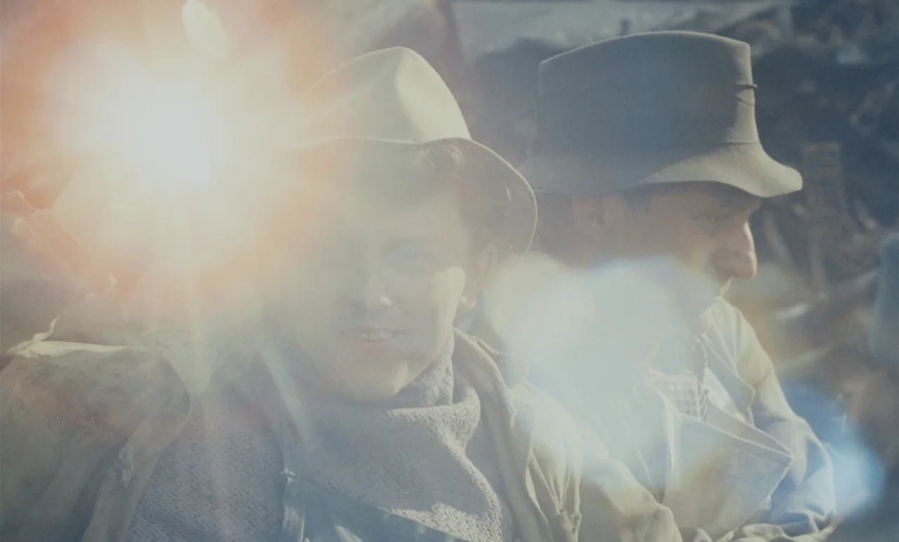 Larks On A String (1969) movie still: medium — Two men wearing fedoras sit closely together in an outdoor setting under bright light. A significant…