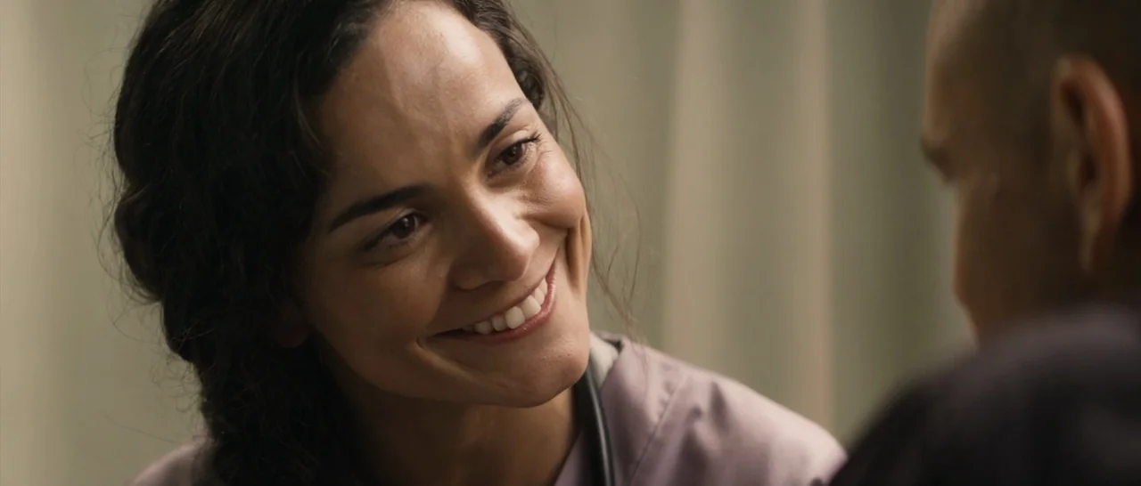 Elysium (2013) movie still: closeup — A hospital room with beige curtains in the blurred background. A smiling nurse looks at a patient in the…