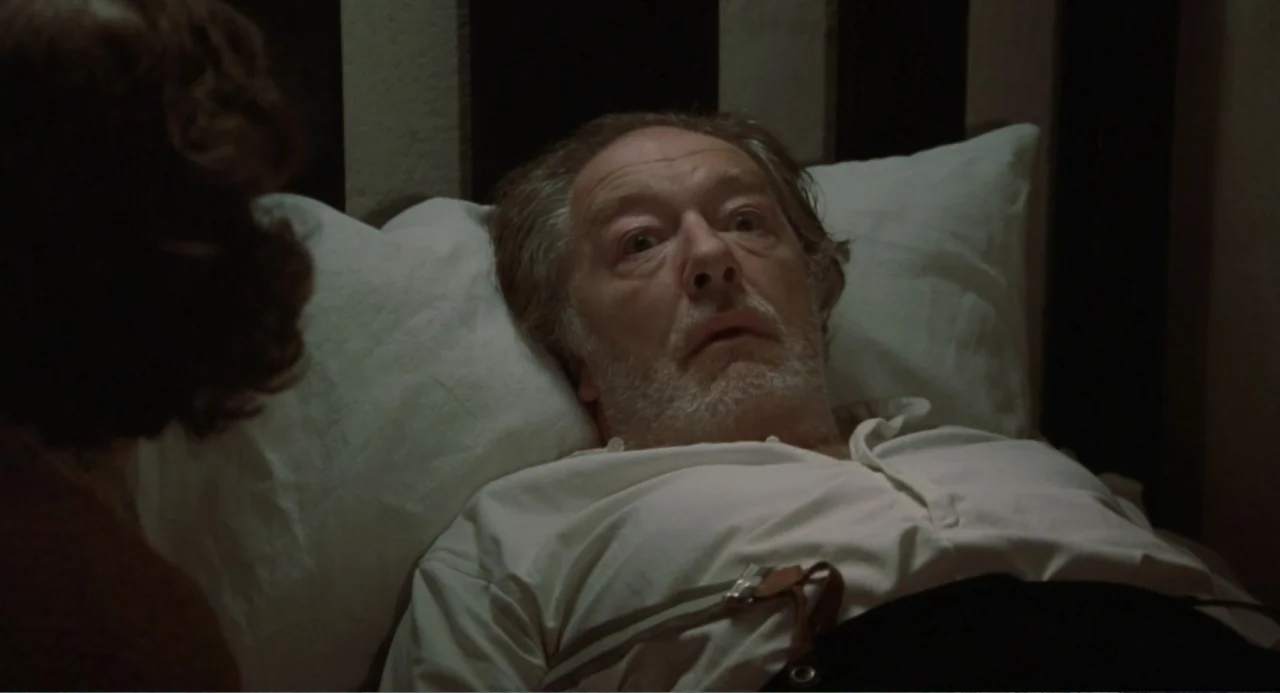 Dancing at Lughnasa (1998) movie still: closeup — A dimly lit room with plain wall panels and white bedding. An elderly man with a beard looks…