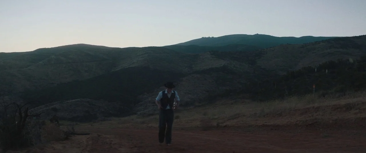 Honey Boy (2019) movie still: wide — A man walks alone on a dusty dirt road at twilight near large hills. The landscape is sparse with dry…
