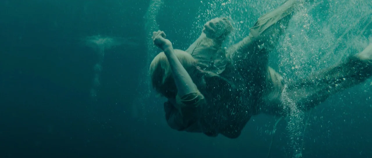 Unbroken (2014) movie still: fullbody — A man sinks vertically into dark open water surrounded by swirling white bubbles. Sunlight penetrates the…