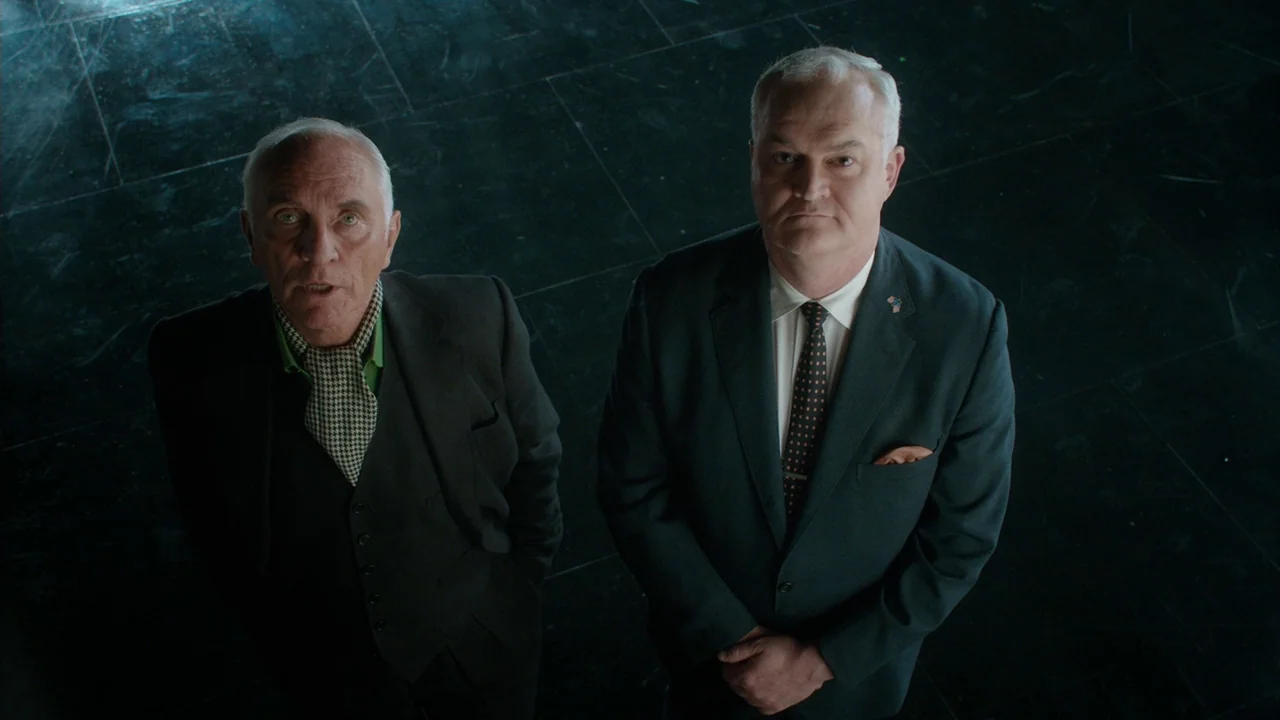 Big Eyes (2014) movie still: medium — Two men stand on a dark, polished stone floor looking straight up. The space is vast and dimly lit with a…