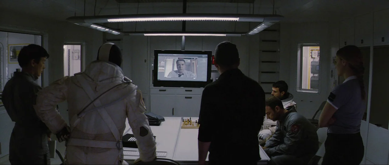 The Last Days On Mars (2013) movie still: wide — A sterile laboratory or briefing room contains a white central table and wall-mounted screen. Five…