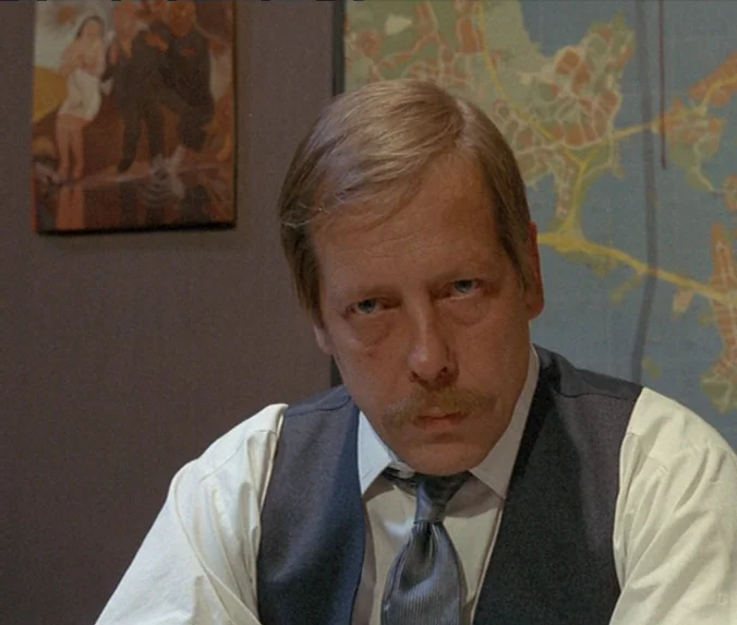 Crime & Punishment (1983) movie still: medium — An interior office space with a wall map and a small framed painting. A middle-aged man with a…