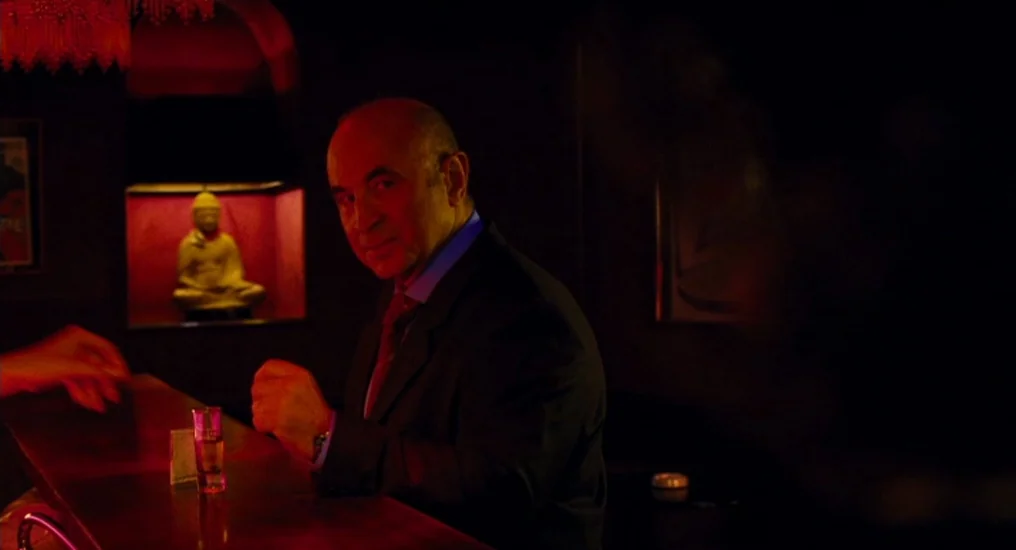 Paris, Je T’Aime (2006) movie still: medium — A dimly lit bar with dark walls and a recessed niche containing a Buddha figure. A man in a suit sits…