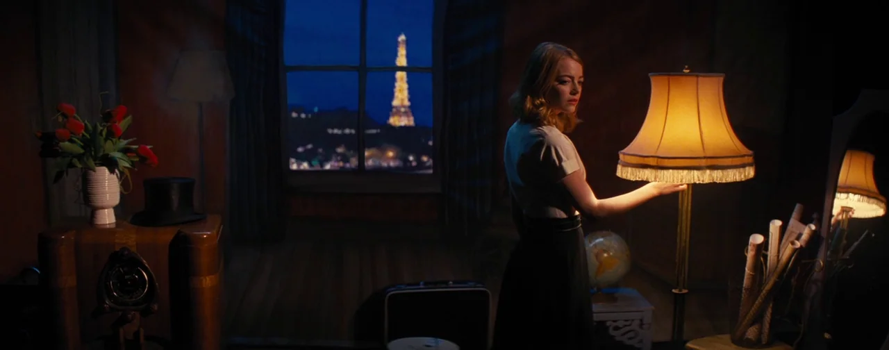 La La Land (2016) movie still: medium — A dimly lit room with vintage furniture and a large window facing the night sky. A woman stands in profile,…