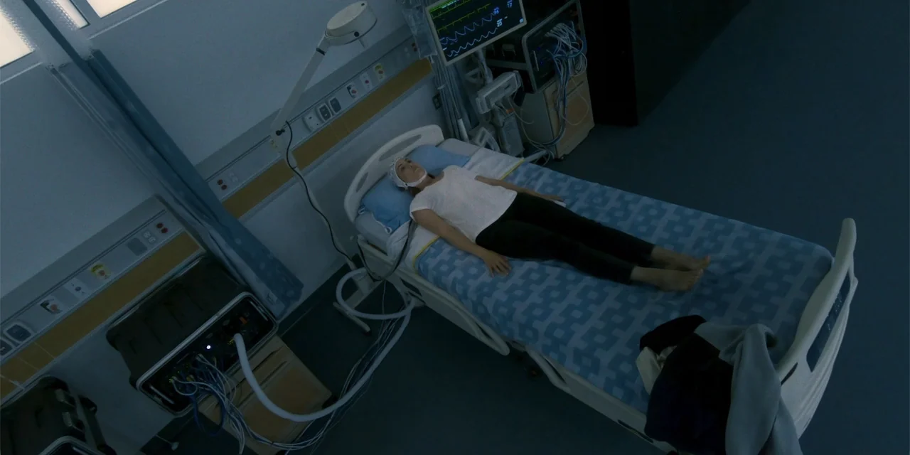 Demonic (2015) movie still: wide — A stark hospital room features a single bed with medical equipment and monitoring displays. A woman lies…