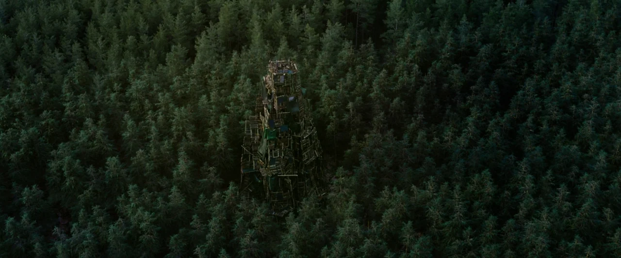 Vesper (2022) movie still: establishing — A dense, uniform pine forest surrounds a tall, conical tower constructed from scavenged wood. The tower…