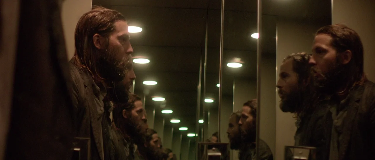 The Invitation (2015) movie still: medium — A man stands inside a small, mirrored elevator illuminated by rows of recessed lights. He observes his…