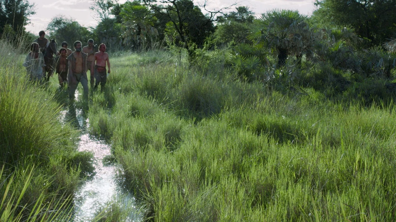 Zama (2017) movie still: wide — A group of men moves through a shallow creek winding through a dense grassy marsh. Palm-like trees and brush fill…
