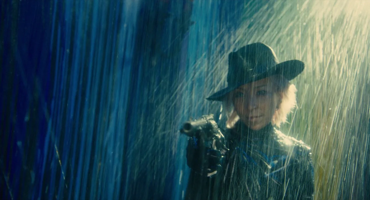 Diner (2019) movie still: medium — An individual stands outdoors during a heavy rainstorm with light rays visible. A woman wearing a hat holds a…