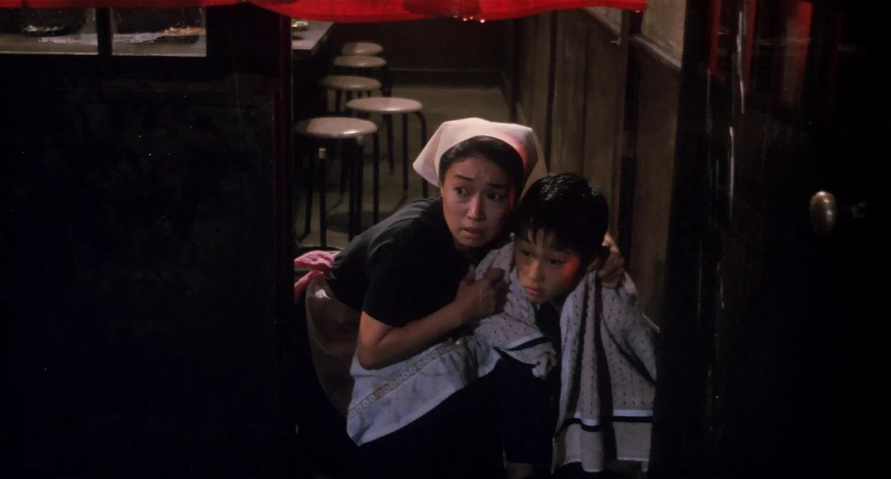 Tampopo (1985) movie still: medium — A dimly lit indoor space featuring multiple circular stools and dark wooden walls. A woman hugs a boy who is…