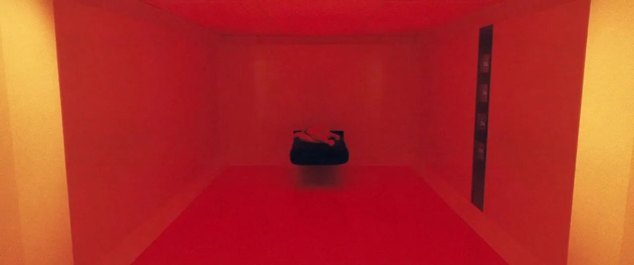 Beyond The Black Rainbow (2010) movie still: wide — A minimalist, rectangular room is bathed in a uniform, deep red light. A person lying on a dark,…