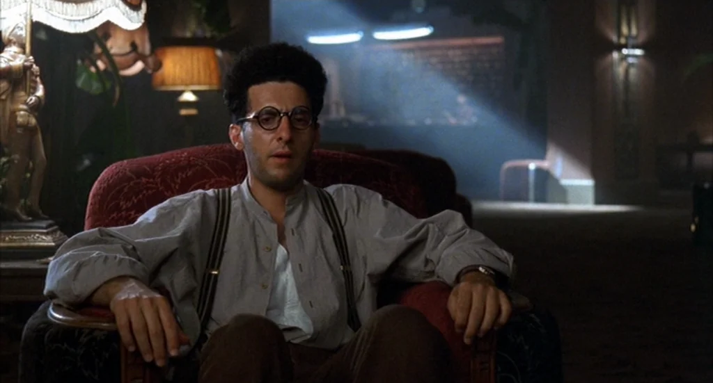 Barton Fink (1991) movie still: medium — A dimly lit, carpeted lobby with decorative lamps and dark furniture. A man sits alone in a red patterned…
