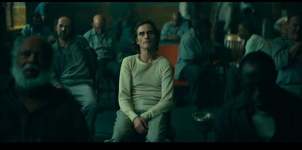 Joker: Folie à Deux (2024) movie still: medium — A dimly lit room filled with men sitting on chairs in a waiting area. A man with shoulder-length…