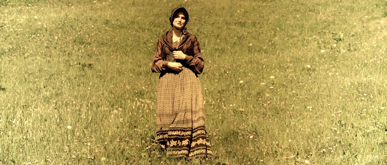 Youth (2015) movie still: fullbody — A vast meadow filled with green grass and small wildflowers under open sky. A young woman in 19th-century dress…