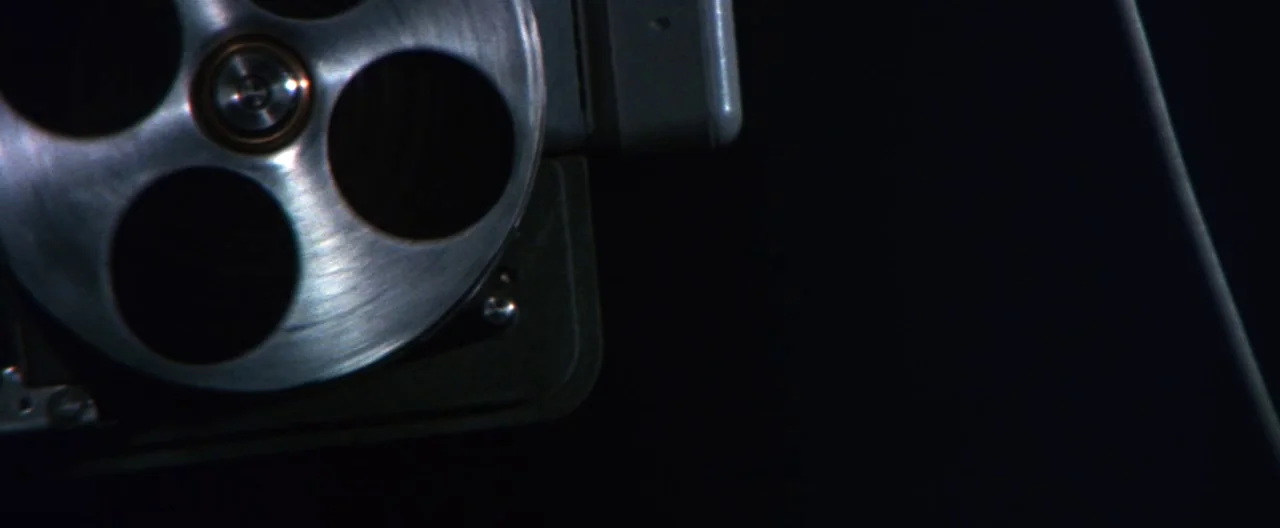 Klute (1971) movie still: closeup — A close view of a metallic film projector reel spins against a dark background. Only a portion of the circular…