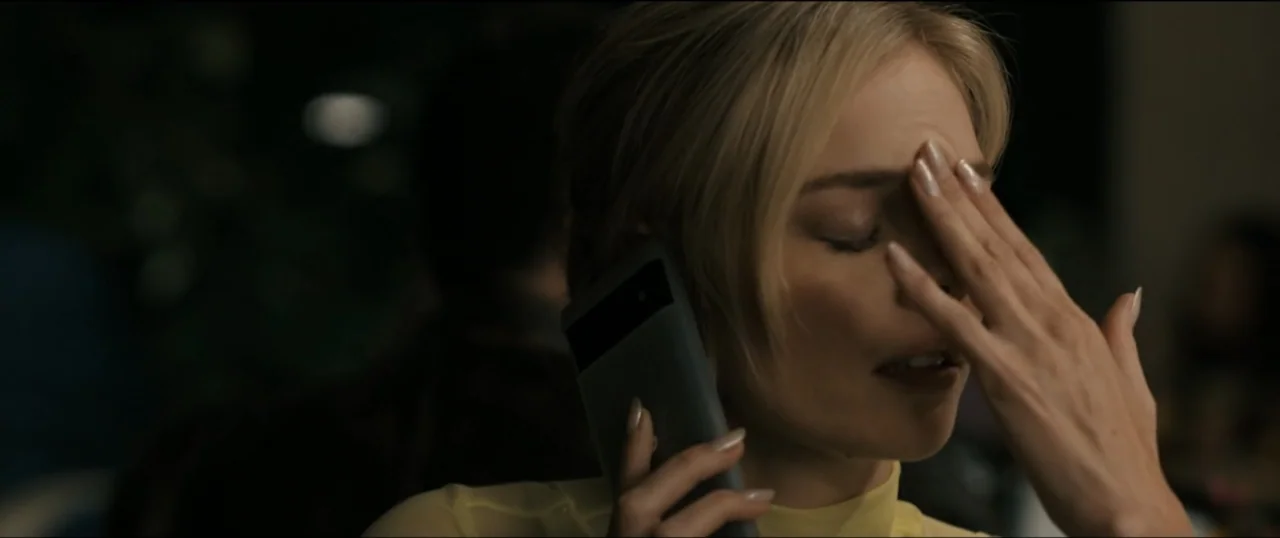 Scream VI (2023) movie still: closeup — A dimly lit interior space contains a person in a yellow garment. A woman holds a smartphone to her ear…