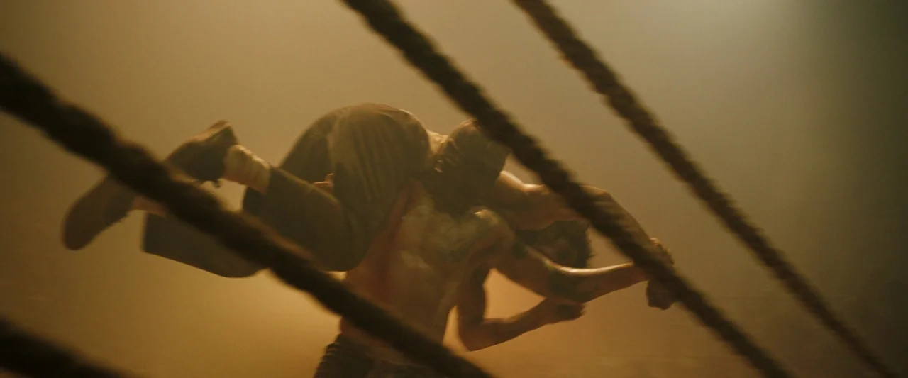 Monkey Man (2024) movie still: medium — A dark indoor space filled with atmospheric haze and overhead warm lighting. Two shirtless muscular men…