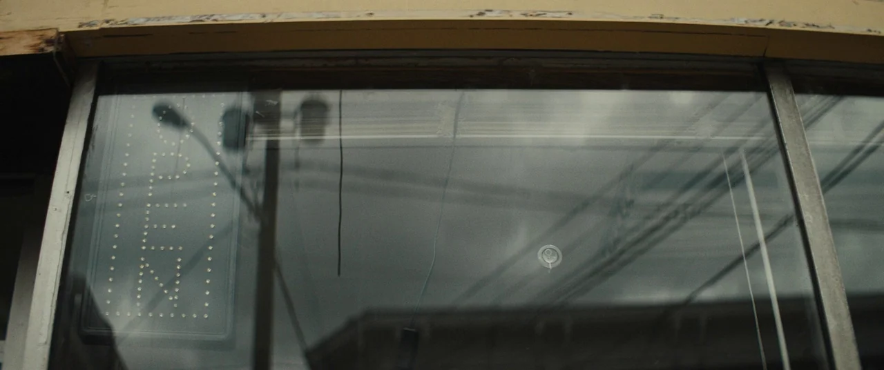 I Think We’re Alone Now (2018) movie still: closeup — A weathered glass window frames a dark storefront with a lit open sign. Power lines and a…