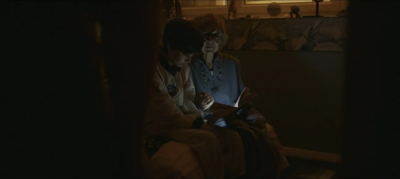 Containment (2015) movie still: medium — A dark bedroom interior features an elderly woman and a child sitting together. The woman reads from an…