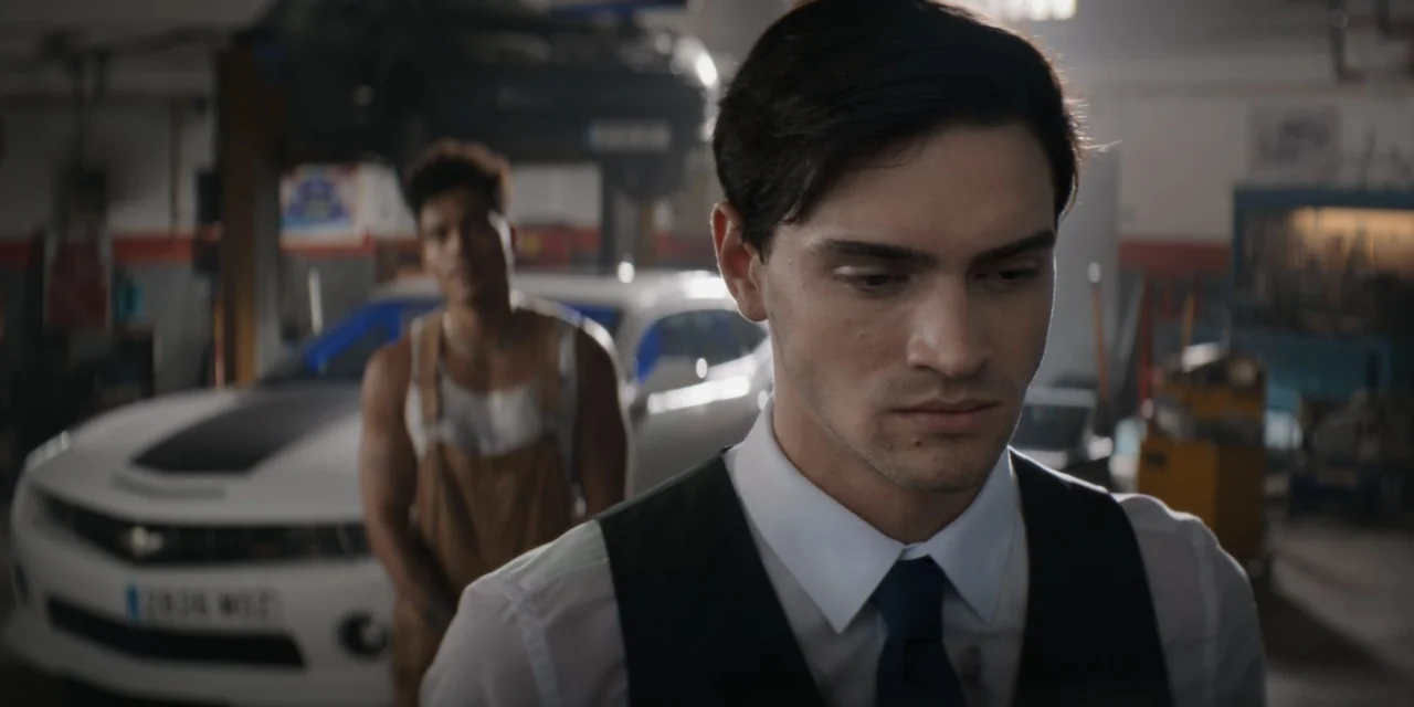 Your Fault (2024) movie still: medium — An auto repair shop interior contains tools, a white car, and a lift. A man in formal attire stands in the…