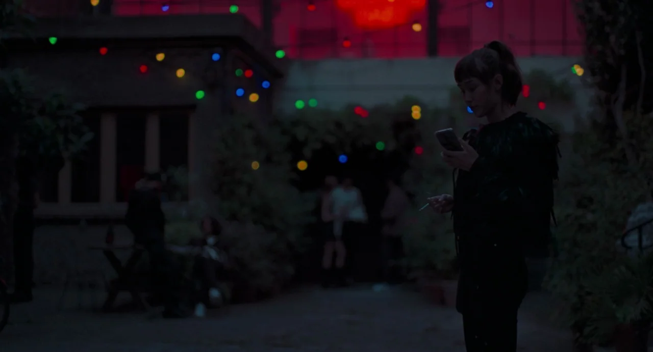 La Abuela (2021) movie still: medium — A dark outdoor courtyard at night features festive string lights draped above figures. A woman stands in the…