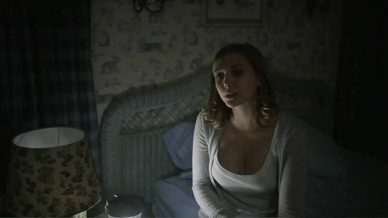 Silent House (2011) movie still: medium — A bedroom at night with patterned wallpaper and a wicker headboard. A woman sits on the bed near a lit lamp.