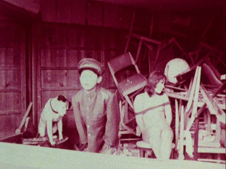 Emperor Tomato Ketchup movie still: medium — A cluttered storage space contains piles of stacked wooden chairs and wooden paneled walls. A young man…