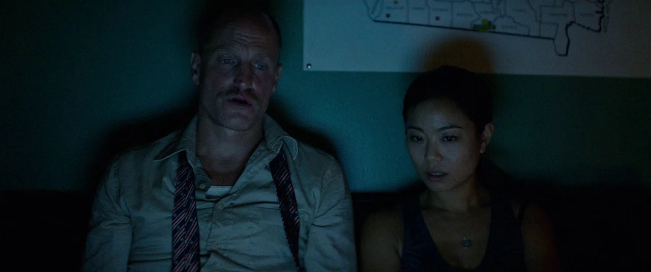 Triple 9 (2016) movie still: medium — A dimly lit room features a wall map hanging behind two seated individuals. A man with a mustache and a woman…