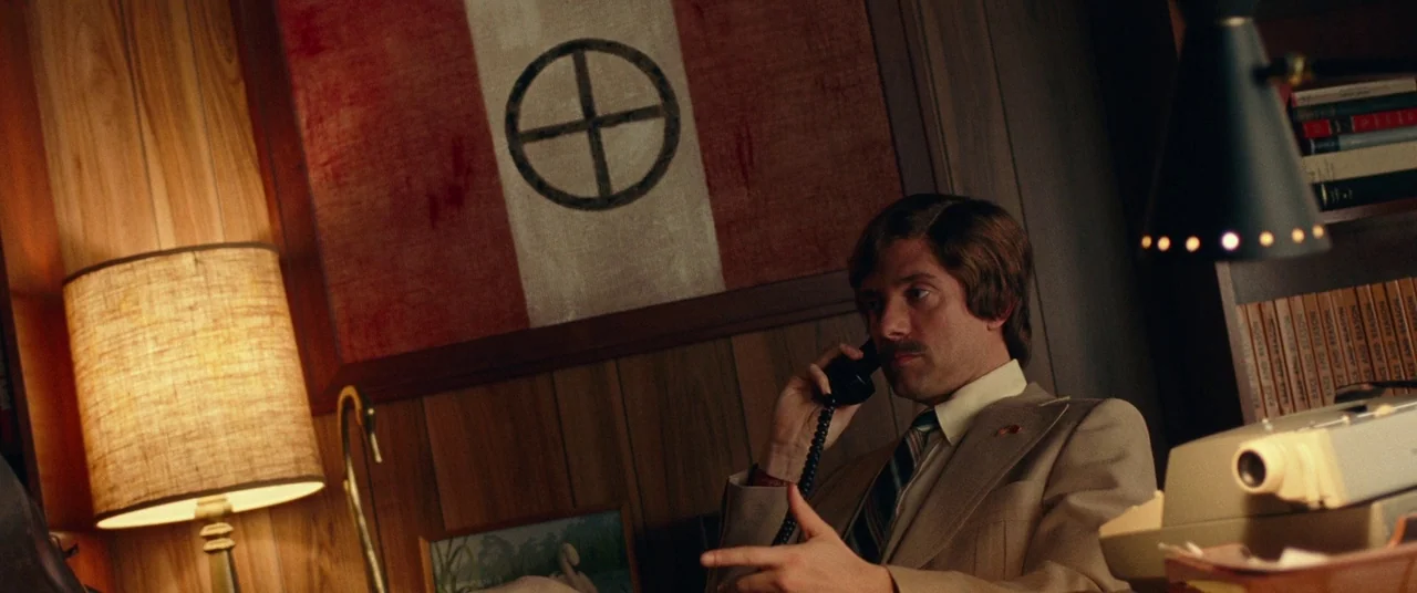 BlacKkKlansman (2018) movie still: medium — A 1970s paneled office features a red and white flag displaying a sun cross symbol. A man with a…