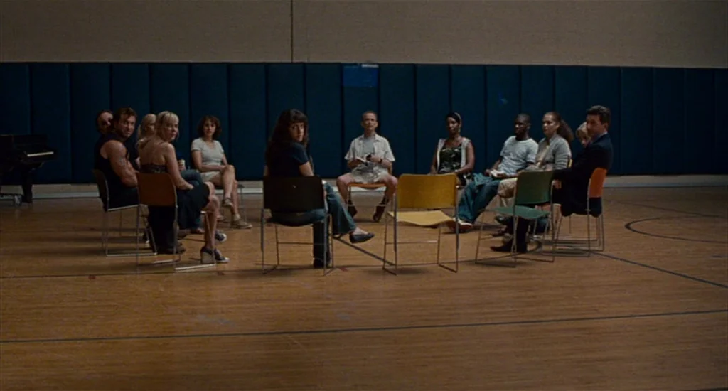 Choke (2008) movie still: wide — A group of people sits in a circle of chairs on a large hardwood court. The walls are covered with dark blue padded…