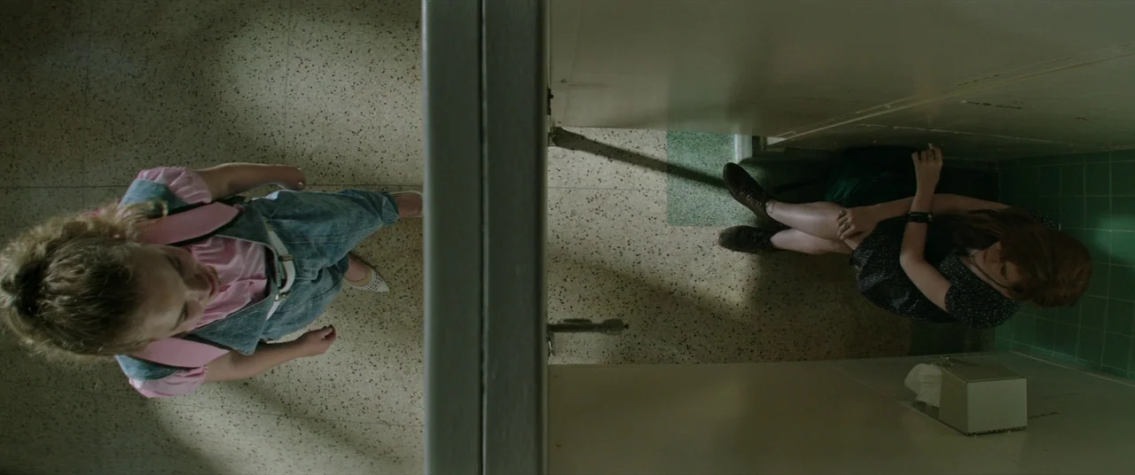 It (2017) movie still: wide — A public restroom interior features speckled tile flooring and divided stalls with metal partitions. One girl in denim…