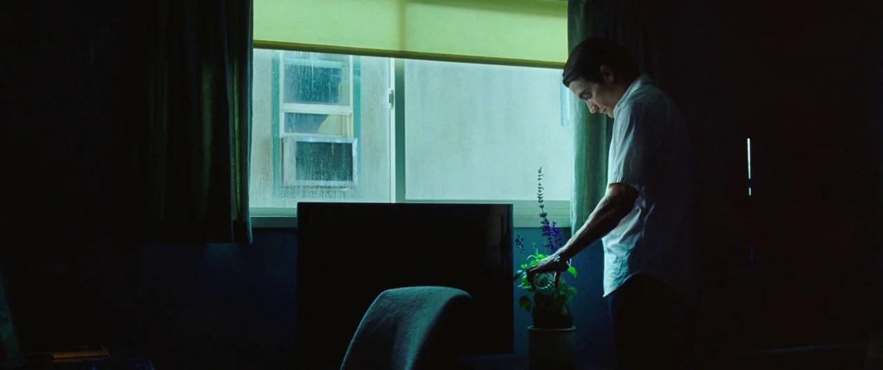 Nightcrawler (2014) movie still: medium — A dark apartment room with a large window looking out onto a neighboring building. A man wearing a white…